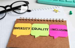 A notebook with three speech bubble sticky notes on it, with the words 'DIVERSITY', 'EQUALITY', and 'INCLUSION' written on them in black ink. The sticky notes are yellow, green, and pink, respectively. In the background, there are a pair of black eyeglasses, a keyboard, a thumbtack, and a pushpin on a checkered paper surface.