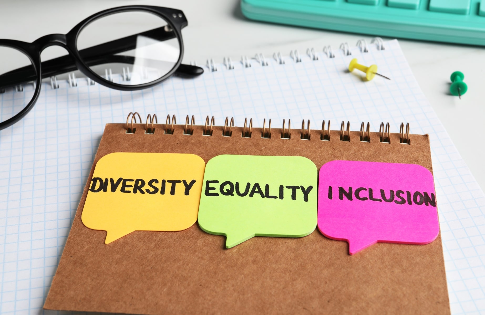 A notebook with three speech bubble sticky notes on it, with the words 'DIVERSITY', 'EQUALITY', and 'INCLUSION' written on them in black ink. The sticky notes are yellow, green, and pink, respectively. In the background, there are a pair of black eyeglasses, a keyboard, a thumbtack, and a pushpin on a checkered paper surface.