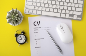 An overhead view of a professional workspace featuring a blank CV template with sections for personal info, contact, profile, work experience, and education. The document lies on a yellow background next to a white keyboard, a white mouse, a green succulent plant in a white pot, and a small black analog alarm clock. The image exemplifies the essentials needed for creating a professional CV.