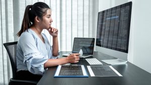 The image shows a woman junior developer sitting at a desk, focused on her work. She is looking at a large computer monitor displaying code, and a laptop with similar code is open next to it. She is holding a piece of paper and appears to be thoughtfully analyzing something. There are documents and a keyboard on the desk, creating a productive, tech-driven atmosphere, possibly related to software development or programming. The setting is a well-lit office with vertical blinds in the background.