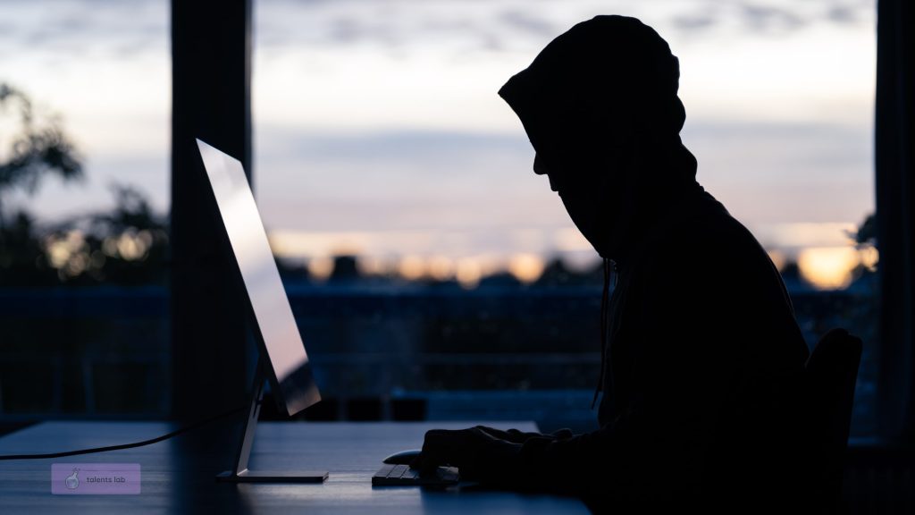 Silhouette of a person in a hoodie using a computer at dusk, representing fake and deep-fake candidates trying to deceive recruiters.