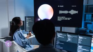 Two professionals in a tech office discussing hiring AI talent, with code on multiple screens and an AI assistant interface displayed on a large monitor.